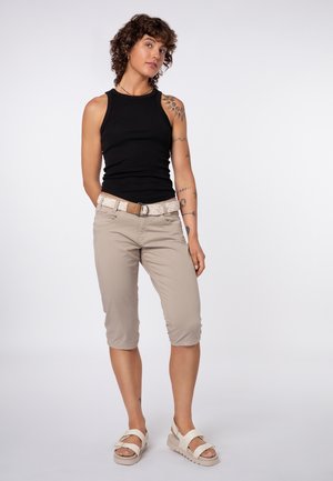 Person with short curly hair wearing a black sleeveless top, beige knee-length pants, white sandals, and a woven belt, standing with hands behind back.