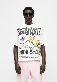 Άνδρας που φοράει μαύρα γυαλιά ηλίου, λευκό υπερμεγέθες μπλουζάκι Adidas Originals με κίτρινη καρτούν πάπια και κείμενο, και ανοιχτό ροζ παντελόνια, στέκεται μπροστά σε λευκό φόντο.