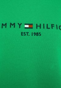 Grön bomull T-shirt med broderad "TOMMY HILFIGER"-logotyp med horisontella f ärgade ränder och "EST. 1985" nedanför. Slät textur.