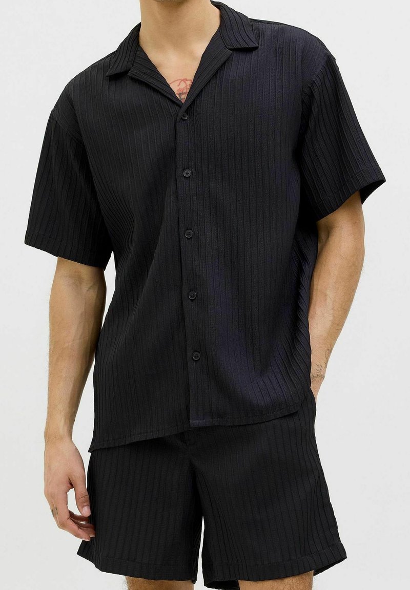 Uomo che indossa una camicia a maniche corte a righe nere con bottoni e pantaloni abbinati, in piedi con una mano in tasca su uno sfondo semplice.