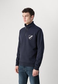 Hanorac cu fermoar navy, cu guler înalt, având un logo brodat alb pe piept și manșete și tiv din țesătură încrețită. Fabricat dintr-un material moale.
