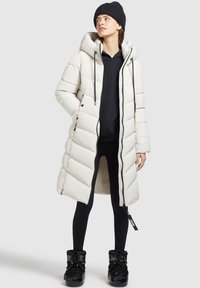 Lang gewatteerde jas in lichtbeige met een capuchon en een ritssluiting. Voorzien van horizontale stiksels, zijzakken en een ontspannen pasvorm. Gecombineerd met zwarte leggings en laarzen.