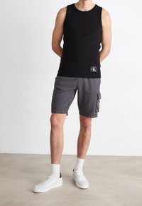 Débardeur noir sans manches avec un logo, associé à un short cargo gris foncé. Baskets blanches avec des accents gris clair et chaussettes blanches. 