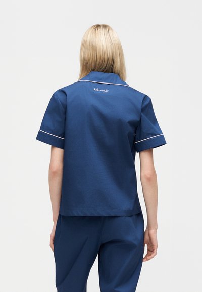 Ξανθός/ή με μπλε σκούρο κοντομάνικο πουκάμισο και παντελόνι που ταιριάζουν, με πλάτη σε ουδέτερο φόντο.