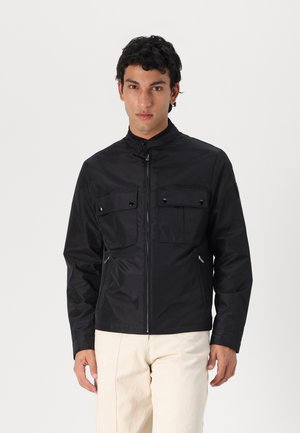 Chaqueta negra con un cuello alto, cremallera frontal y dos bolsillos en el pecho. Presenta una textura suave y un diseño discreto. Se lleva con pantalones de color claro.