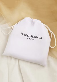 Pochette blanche à cordon avec l'inscription "ISABEL BERNARD PARIS" imprimée en noir, placée sur un fond en tissu beige clair et crème.