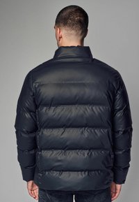 Chaqueta acolchada negra con un alto cuello, que presenta secciones acolchadas horizontales y una textura brillante y lisa. Capucha adjunta visible.