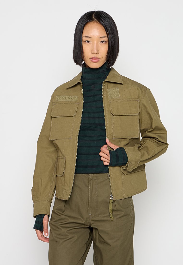 Olivgrüne Jacke mit vier Fronttaschen, einem Reißverschluss und einem Kragen. Getragen über einem dunkelgrünen gerippten Pullover.