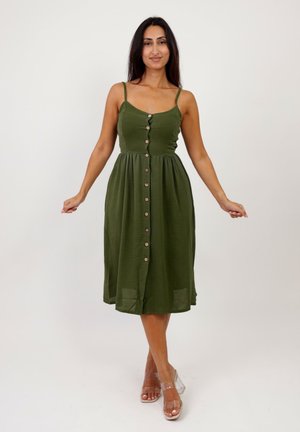 Robe d'été en tissu vert olive avec des fines bretelles, un corsage ajusté et une jupe évasée ornée de boutons marron sur le devant. Talons transparents.