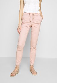 Žena v svetle růžových slim fit kalhotách s pásem na stažení a plochými sandály se zubatými pásky stojí na jednobarevném pozadí.
