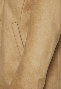 Tissu beige clair à texture douce en gros plan, montrant une poche latérale avec des coutures nettes et des coutures minimales. Design simple avec une finition lisse.