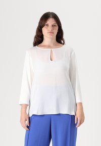 Witte, semi-doorzichtige blouse met een ronde hals en sleutelgatdetail, lange mouwen en een ontspannen pasvorm; gedragen met blauwe wijde broek.