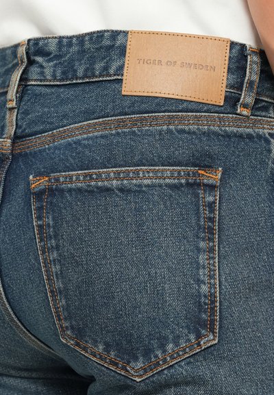 Jeans en denim avec un lavage bleu foncé, arborant une étiquette en cuir portant l'inscription "TIGER OF SWEDEN." Comprend des coutures orange et deux poches arrière.