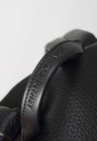 Sac en cuir noir avec une surface texturée, doté d'une poignée élégante estampillée "ARMANI EXCHANGE." Détails de couture visibles.