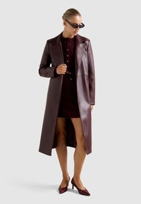 Μακρύ παλτό από ψεύτικο δέρμα μπορντό με γιακά με σχισμή, ελαφρώς εφαρμοστή γραμμή και λεπτομέρειες με κουμπιά, σε συνδυασμό με ασορτί φόρεμα και μυτερές γόβες.