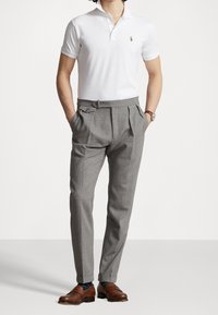 Man med vit polo-shirt, grå veckade byxor, bruna loafers och klocka, står med händerna i fickorna mot en enkel bakgrund.