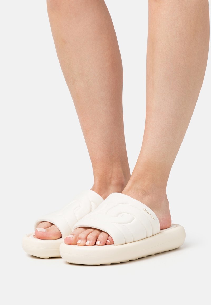 Sandales à glissière blanches avec dessus matelassé, bout rond et semelle épaisse et rembourrée. Détail discret du logo sur le côté. Texture lisse dans l'ensemble.