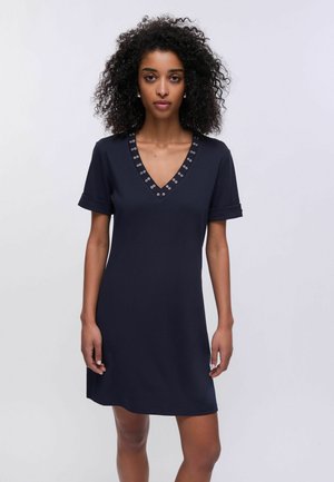 Donna con capelli ricci che indossa un vestito blu navy a maniche corte con scollo a V e dettagli in occhiello metallico, in piedi davanti a uno sfondo semplice.