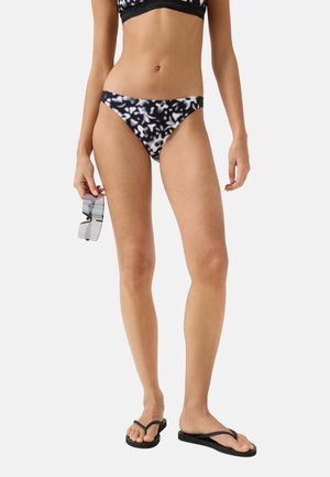 Frau trägt schwarzen und weißen gemusterten Bikinihöschen und schwarze Flip-Flops, hält eine klare Sonnenbrille vor weißem Hintergrund.