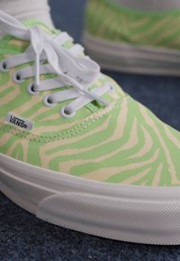 VANS sneakers with a light green zebra pattern on canvas material, white laces, and a white rubber sole. Positioned on a textured surface.