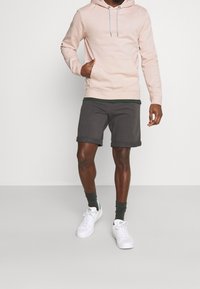 Sudadera rosa claro con bolsillo frontal y cordones, combinada con pantalones cortos grises oscuros con dobladillos enrollados y zapatillas blancas.