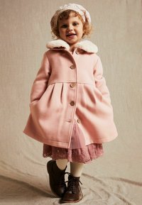 Lyserød uldfrakke med faux fur-krave, træknapper og plisseret design. Båret over en lagdelt lyserød kjole med blondekant og brune støvler.