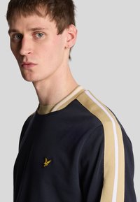Sweat-shirt marine avec des rayures beige et blanches sur les épaules, col rond, et un logo jaune brodé sur la poitrine. Tissu doux et texturé.