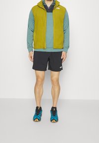 Fleece vesta v olivově zelené barvě přes tyrkysové tričko s dlouhým rukávem, v kombinaci s černými šortkami a modro-černými sportovními botami.