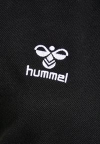 Czarna tkanina z białym haftowanym logo pszczoły, z nazwą marki "hummel" poniżej. Tekstura wydaje się gładka i gęsta.