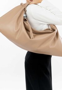 Person der holder en stor beige læder skuldertaske over hvid bluse og sorte bukser mod en hvid baggrund.