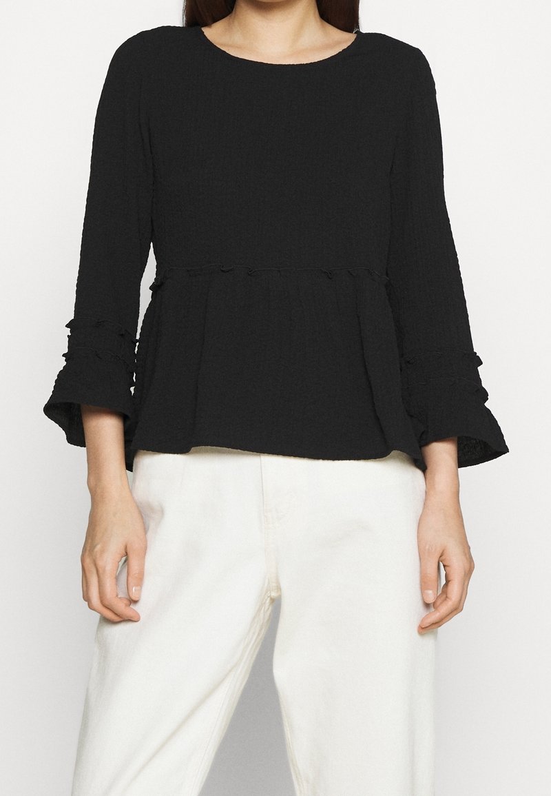 Femme portant un chemisier noir texturé à manches longues avec des poignets évasés et un pantalon blanc à jambes larges, debout devant un fond uni.