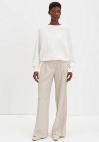 Maglione bianco a maniche lunghe abbinato a pantaloni larghi beige. Il modello indossa pantaloni chiari plissettati e scarpe cremosi a trama.