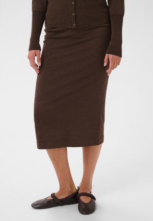 Femme portant un cardigan marron boutonné et une jupe midi marron assortie avec des chaussures plates noires sur un fond blanc uni.