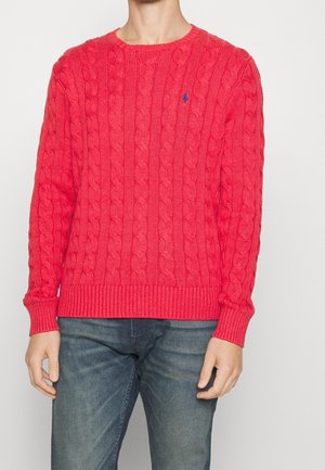 Man wearing a bright red cable-knit sweater with long sleeves and blue jeans, standing against a plain light background.