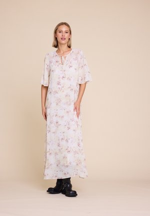 Kvinne iført en lang, lys blomsterkjole med albue-length ermer, kombinert med svarte støvler med spenne, stående mot en beige bakgrunn.