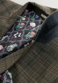 Ruskea ruudullinen bleiseri, jossa on tekstuurinen kangas ja tumman kukallinen vuori purppuran, vihreän ja valkoisen sävyissä.