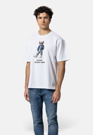 Jonge man met een wit T-shirt met een afbeelding van een beer in een varsityjack en spijkerbroek met verfspatten en scheuren.