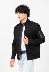 Zwarte zip-up jack van gestructureerde stof met een staande kraag, een ritszak op de mouw en een geborduurd logo op de borst.