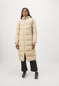 Lunga giacca a vento beige con cappuccio, caratterizzata da una chiusura con zip verticale, design trapuntato e polsini elastici, abbinata a pantaloni e scarpe neri.