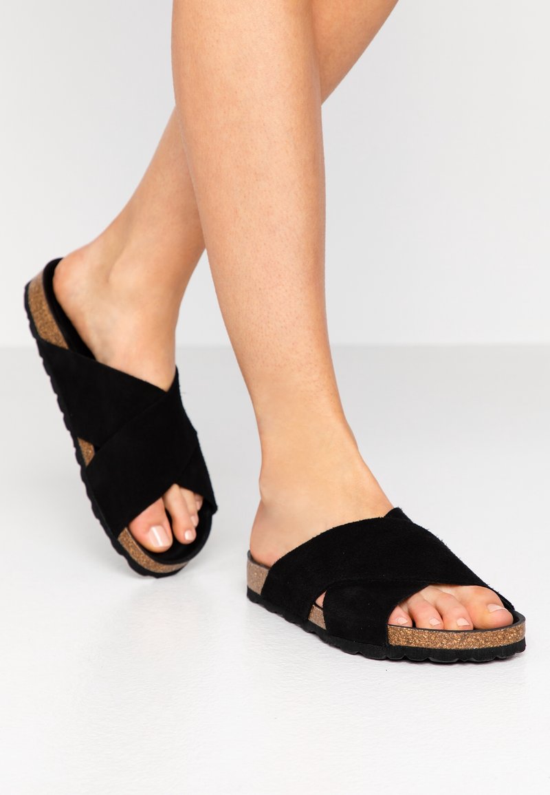 Sandales à glissière en daim noir avec design à lanières croisées, semelle plate en liège et bout ouvert. Texture douce, avec une finition lisse sur la semelle.