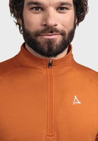 Oranges, langärmliges Sportoberteil mit einem halben Reißverschluss und strukturiertem Stoff. Weißes Logo-Detail auf der Brust. Nahaufnahme.