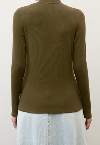 Une personne portant un haut à manches longues côtelé vert olive et une jupe en denim bleu clair, vue de dos contre un fond uni.