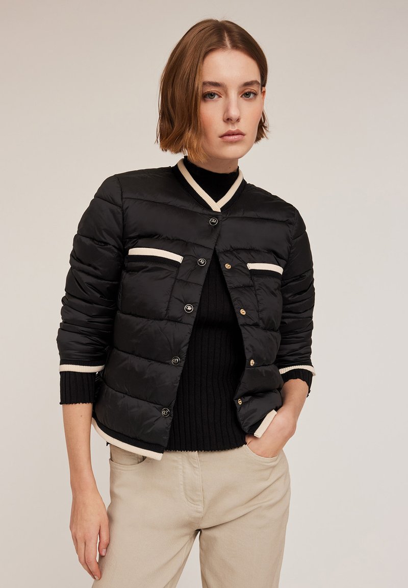 Giubbotto imbottito nero con un design a nido d'ape, dettagli color crema, bottoni a pressione e polsini e colletto a coste, indossato sopra un maglione nero.