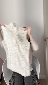 Άτομο που κάθεται σε λευκή καρέκλα κρατώντας και ξεδιπλώνοντας ένα πλεκτό πουλόβερ σε κρεμ χρώμα μέσα σε εσωτερικό χώρο, με απλή λευκή επιφάνεια τοίχου στο φόντο.