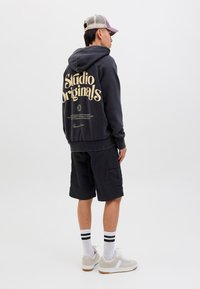 Černá mikina s kapucí s nápisem "Studio Originals" vytištěným v krémové barvě, doplněná černými kraťasy. Doplňky zahrnují růžovou a šedou kšiltovku, bílé ponožky s černými pruhy a šedé tenisky.