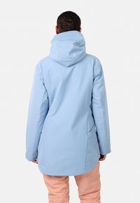 Helleblaue wasserdichte Jacke mit Kapuze, langen Ärmeln und mittellanger Schnittform. Sie verfügt über ein Logo auf dem Rücken und eine glatte Textur. Kombiniert mit pinken Hosen.