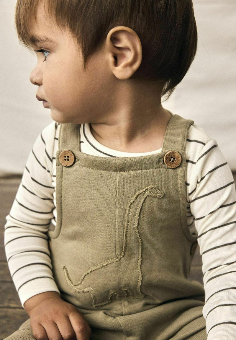 Ternura con un overol de un suave verde oliva adornado con un bordado de dinosaurio. Presenta detalles de botones de madera y una camiseta de rayas de manga larga debajo.