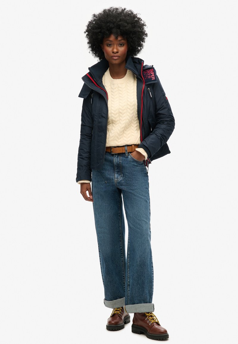 Marineblauwe pufferjack met rode accenten, beige kabelgebreide trui, lichtblauwe high-waisted jeans en bruine veterschoenen. Textuurvolle, casual outfit.