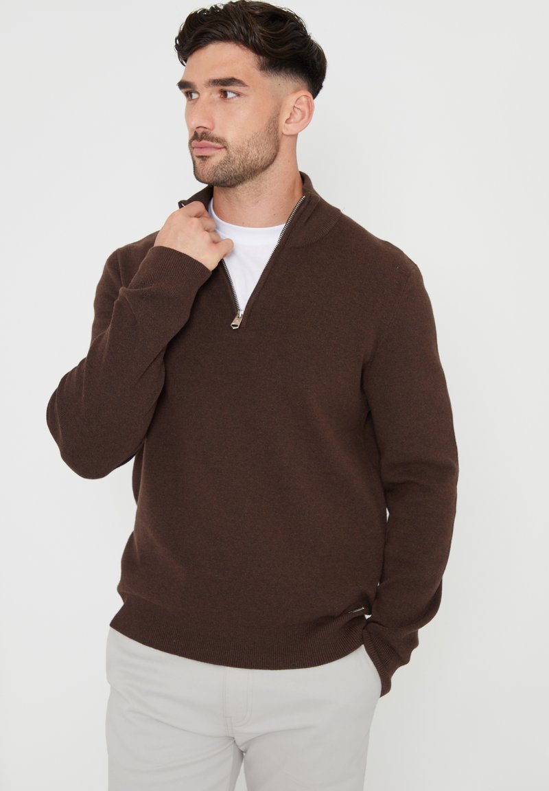Brauner Zip-Kragen-Pullover aus gestricktem Material mit ribbeliger Saum- und Bündchenverarbeitung. Über einem weißen T-Shirt getragen, mit einem silbernen Reißverschluss-Detail.