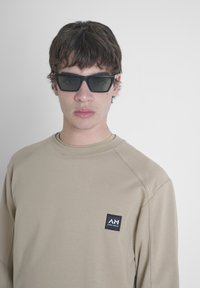 Bež dukserica s okruglim izrezom i raglan rukavima, s crnim logotipom na lijevoj strani. Model nosi tamne naočale.